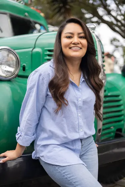 young person with long hair smiling in front of a vintage car, relaxed lifestyle hacinedo tourism, background with sunlight, beauty and casual fashion of latin woman
