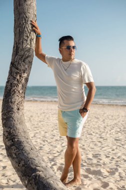 Tatilin tadını çıkarıyorum, güneşli bir günde güneş gözlüklü, açık renk elbiseli genç Latin bir adam plajda yürüyor, rahat bir yaşam tarzı, erkek modası.