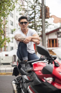 Şehirde sağlıklı vücutlu, spor motosikletinin yanında duran güneş gözlüğü takan, yaz aylarında manken portresi giyen çekici bir genç adam bekliyor.