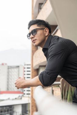 Siyah ve resmi giyinmiş modern bir insan, balkona yaslanırken şehrin, güzelliğin ve modanın tadını çıkarıyor, Latin adam.