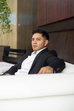 Siyah takım elbiseli, kısa saçlı, zarif ve iş adamı bir gencin portresi. Kanepede, ofis içinde dinleniyor.