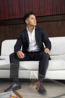 Bir kanepede dinlenen, kısa saçlı, siyah şık takım elbiseli, beyaz gömlekli, profesyonel ve yaşam tarzının portresi, bekleyen genç bir adam.