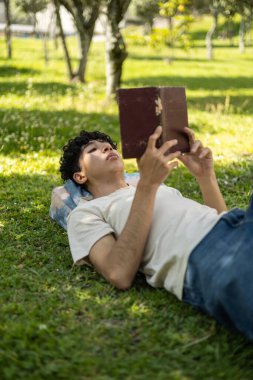 Parktaki çimlerin üzerinde uzanmış kitap okuyan bir çocuk. Rahatlama, okuma, açık hava aktiviteleri ve huzurlu anlar için idealdir..
