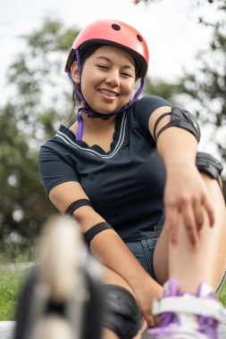 Güzel bir genç kız yeni patenleriyle, kaskıyla, dizlikleriyle ve dirseklikleriyle poz verirken kendine güvenerek gülümsüyor. Eğlenceli ve şık bir hava yayıyor..
