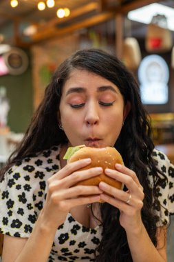 Genç bir kadın iki eliyle sulu bir burger tutuyor. Lezzetli bir ilk ısırık umuduyla gözlerini kapıyor..