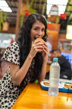 Tek renkli elbiseli moda bir kadın lüks bir restoranda esnaf hamburgerini ısırıyor. Önde limonlu su. Otantik samimi an.