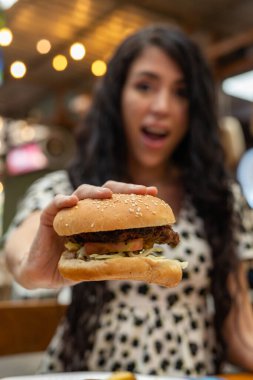 Bir kadın hamburgerini iki eliyle tutar, huşu ve şaşkınlıkla bakar, neşeli ve iştah açıcı bir yemek anını yakalar..