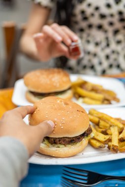 Bir kadın kızarmış patatese ketçap ekliyor. Yanında da hamburger var. Sıradan bir akşam yemeğinin keyfini çıkarıyor..