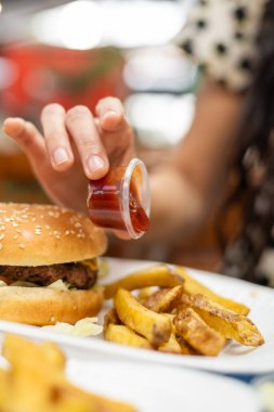 Siyah & beyaz elbiseli şık bir kadın restoran masasındaki çıtır patateslerin üzerine ketçap sıkıyor. Hamburger ve içecekle otantik fast food keyfi.