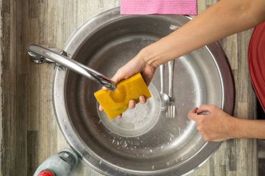 Bir kadının eli suyun altındaki süngeri ıslatır, aşağıdaki lavaboda bulaşıkları yıkamaya hazır, temiz, modern bir mutfak alanında..