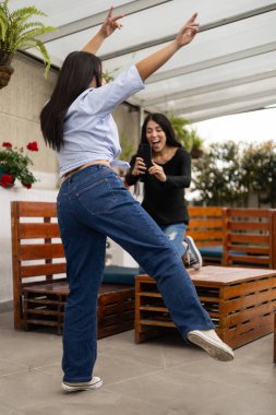 İki kadın, ahşap bir verandada masalarla fotoğraf çekerken, keyifli anları paylaşıyor. Arkadaşlıkları harmanlıyor, açık havada rahatlıyor ve doğal güneş ışığı altında gündelik tarzlarını sergiliyor..