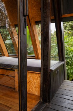Açılı cam pencerelerle çevrili rahat bir bank, modern ahşap tasarımı tropikal doğayla harmanlayan yeşilliğin huzurlu bir görüntüsünü sunar..