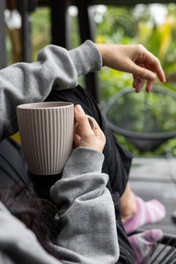 Bir kadın, bir bacağı esnemiş ve diğeri katlanmış bir şekilde, kupasını tutarken tahta bir duvara yaslanmış olarak rahatlar. Huzurlu bir şekilde orman manzarasına bakıyor..