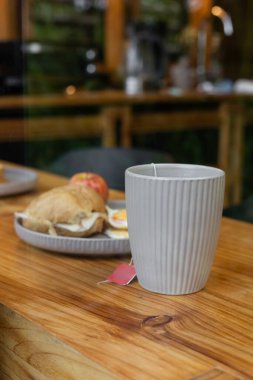 Sıcak bir kahve bardağının yanında içinde peynirli sandviç ve taze elma olan bir tabağın yanında, ahşap bir masaya kurulmuş, samimi ve davetkar bir kahvaltı atmosferini çağrıştıran