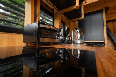 Şık bir ahşap glamping mutfağına yerleştirilmiş küçük bir elektrikli ekmek fırınının yakın çekimi. Modern aletler ve doğal malzemelerin zarif karışımı sıcak ve rahat bir atmosfer yaratır..