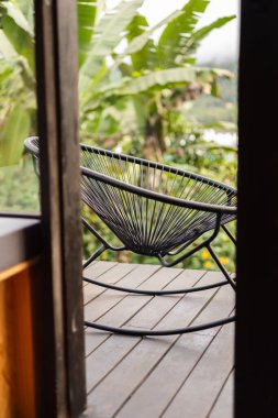 Yuvarlak örülmüş bir sandalye tropikal ormanlarla çevrili kırsal bir glamping kulübesinin balkonunda duruyor. Bir muz bitkisi bu huzurlu açık havaya bereketli bir dokunuş katıyor..
