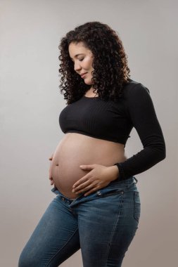Işıl ışıl hamile bir kadın gururla durur, gururla bebeğini gösterirken şık bir siyah bluz ve kot pantolon giyer, annelikte güçlü ve güzel bir vücut bulur..