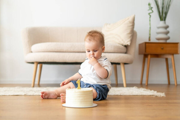 A baby is sitting on the floor next to a colorful cake, looking curiously at the delicious treat in front of them.