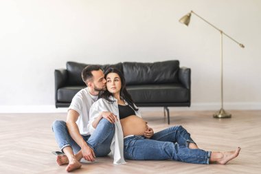 Hamile bir çift oturma odalarının zemininde oturmuş ev dekoruyla çevrili bir şekilde sohbet ediyorlar..