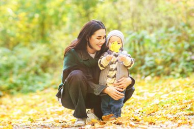 Anne ve çocuğu düşmüş yapraklarla ormanda oynuyorlar. Çocuk, hem gülüp hem de eğlenirken sarı yaprağı gözlerinin önünde tutuyor..