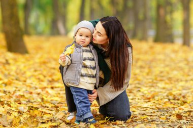 Bir anne, sarı bir yaprak tutan oğluna bakmak için diz çöker. İkisi de sonbahar yapraklarıyla kaplı bir parkta duruyorlar..