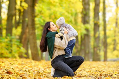 Bir anne, sarı yapraklardan oluşan bir ormanda diz çöker ve oğlunu yanağından öpmek için uzanırken kucağına alır..