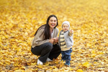 Bir anne, düşen sonbahar yapraklarıyla kaplı bir parkta oynarken oğluna gülümser. Çocuk heyecanla uzaktaki bir şeyi işaret ediyor..