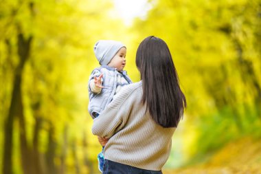 Bir anne bebeğini kucağında tutup ona gülümsüyor. Arka plan, sarı ve yeşil yapraklı bir ormanın bulanık görüntüsüdür..