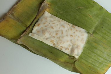 Bir dilim tempeh ya da tempe, mayalanmış soya fasulyesi, muz yapraklarına sarılmış, beyaz arka planda. Pişmemiş ya da çiğ tempeh. Düz ya da üst görünüm