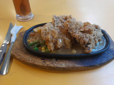 Elektrikli ocakta çıtır biftek, mantar sosu ve sebzelerle servis edilmiş, yanında da metal çatal bıçak ve buzlu çay.