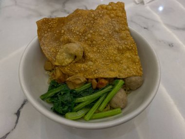 Endonezya köftesi, kızarmış hamur köftesi, sebze olarak yeşil hardal yaprağı, et suyu olmadan servis edilir. Beyaz kâse, beyaz mermer masa, üst manzara.