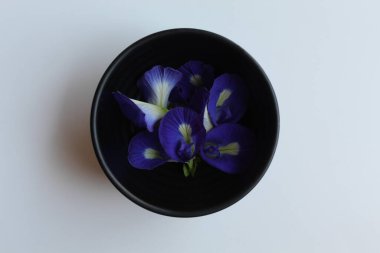 Pile of purple flowers of Clitoria ternatea, or butterfly pea flower or bunga telang, in black bowl, isolated on white background, flat lay or top view clipart