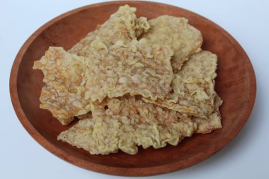 Keripik tempo veya tempeh krakerleri, kare şeklinde. İnce dilimler, un hamuruyla kızartılmış tempeh. Tahta tabakta, beyaz arka planda izole edilmiş.