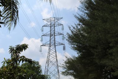 Yüksek voltajlı elektrik kuleleri ve hatları, ağaçların arasında, bulutlu mavi gökyüzü arkaplanı