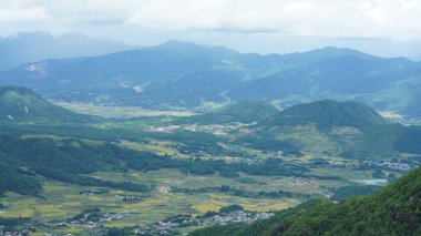 Arkaplanı bulutlu gökyüzü olan dağların arasındaki vadide bulunan sarı pirinç tarlası manzarası.