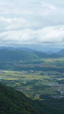 Arkaplanı bulutlu gökyüzü olan dağların arasındaki vadide bulunan sarı pirinç tarlası manzarası.