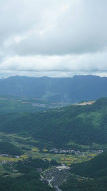 Arkaplanı bulutlu gökyüzü olan dağların arasındaki vadide bulunan sarı pirinç tarlası manzarası.