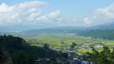 Arkaplanı bulutlu gökyüzü olan dağların arasındaki vadide bulunan sarı pirinç tarlası manzarası.