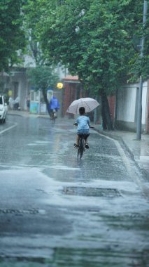 Küçük bir çocuk yağmurlu bir günde şemsiyeyle sokakta bisiklet sürüyor.