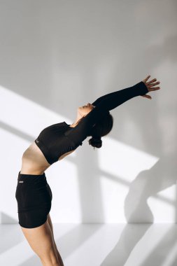 Genç bir kadın yoga antrenmanı sırasında yukarı doğru selam duruşu yaparken yakalanır, kollarını başının üstüne uzatarak güneşli bir odada dikilir. Siyah spor kıyafetleri giymiş, odak noktasını somutlaştırıyor.