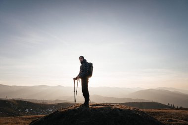 Güneş doğarken bir dağ zirvesinde tek başına duran bir yürüyüşçü, yürüyüş direkleri ve sırt çantasıyla donatılmış, dağ sırasının geniş manzarasının keyfini çıkarıyor..