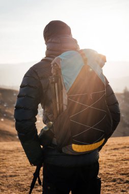 Sırt çantası ve yürüyüş direkleri olan, gün doğarken bir dağın tepesinde duran, altın ışıkla yıkanmış bir yürüyüşçü..