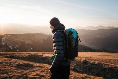 Bir yürüyüşçü gün doğumunda bir dağ yolu boyunca yürür, bir sırt çantası ve yürüyüş direkleri taşır. Yükselen güneşin altın ışığı, doğayla olan bağlantıyı vurgulayan, sıcak ve sakin bir atmosfer yaratır..