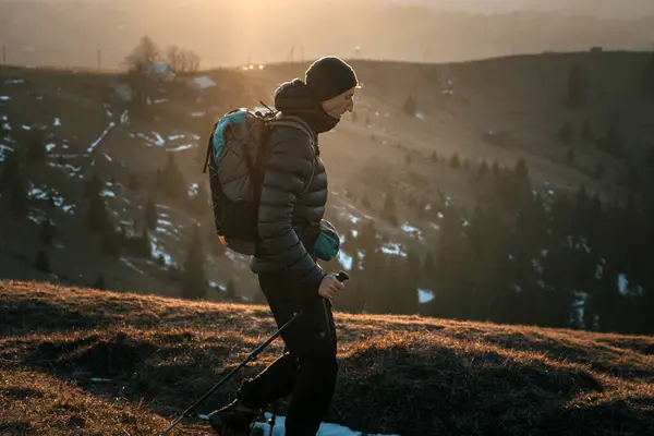 stock image A hiker walks along a mountain trail at sunrise, carrying a backpack and trekking poles. The golden light of the rising sun creates a warm, serene atmosphere, emphasizing the connection with nature.