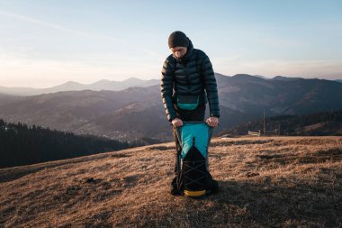 Aşağı ceket giyen bir yürüyüşçü gün batımında manzaralı bir dağ sırtında sırt çantası taşır. Huzurlu manzara ve uzak sıradağlar sakin bir açık hava ortamı yaratır..