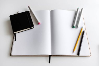 Kalemler, kalemler ve beyaz bir masaya yerleştirilmiş daha küçük siyah bir günlük ile açık bir defter. Minimalist bir çalışma alanında yaratıcılık, üretkenlik ve günlük gibi konular için ideal.