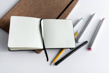 Kalemler, kalemler ve beyaz bir masaya yerleştirilmiş daha küçük siyah bir günlük ile açık bir defter. Minimalist bir çalışma alanında yaratıcılık, üretkenlik ve günlük gibi konular için ideal.