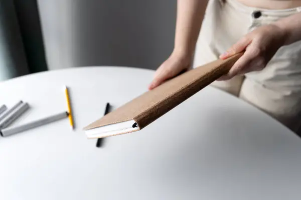 stock image Close-up image of a woman's hands holding a cork-covered sketchbook on a white table with drawing tools scattered. Perfect for art, creativity, and stationery-related themes.