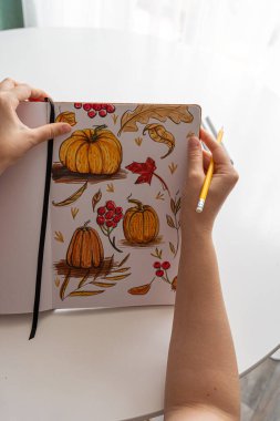 Bir insan, sonbahar temalı resimlerini balkabaklarıyla çizer ve sarı bir kalem kullanarak eskiz defterine bırakır. Yaratıcı ifadeleri vurgulayan çeşitli işaretler etrafa saçılmış durumda..