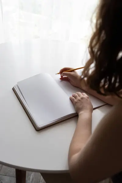 stock image A woman holding a pencil over a blank notebook while sitting at a table near a window. The scene symbolizes creativity, new beginnings, and inspiration in a calm and peaceful environment.